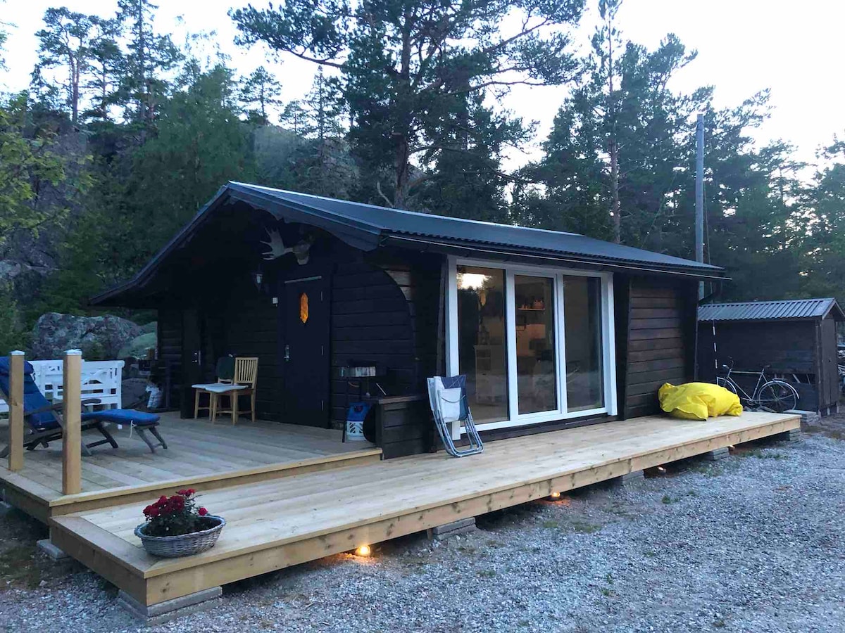 A wooden cabin is shown on a spacious deck, surrounded by trees. Glass doors provide access to the interior, while outdoor seating includes chairs and a small table. A vibrant yellow object rests on the deck, adding a touch of color to the natural setting.