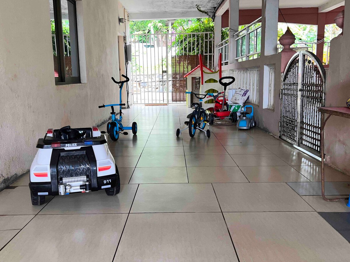 A covered outdoor area features smooth tiled flooring. Several children's ride-on toys, including a police car and tricycles, are positioned on one side, creating a playful atmosphere. Natural light brightens the space from nearby openings while a gate adds security.