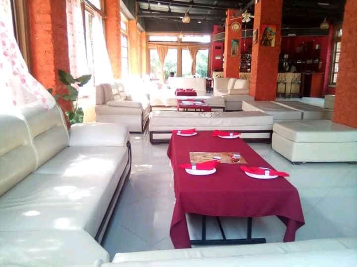 A spacious lounge area is furnished with light-colored seating arranged around low tables covered with red tablecloths. Natural light filters through large windows, illuminating the open space which features a casual and welcoming atmosphere.