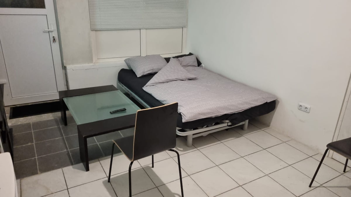 A simple studio space is presented with a cozy sleeping area featuring a neatly made bed and two pillows. A glass table accompanies one black chair, providing a functional workspace. Natural light filters through a window, adding to the minimalistic and inviting ambiance.