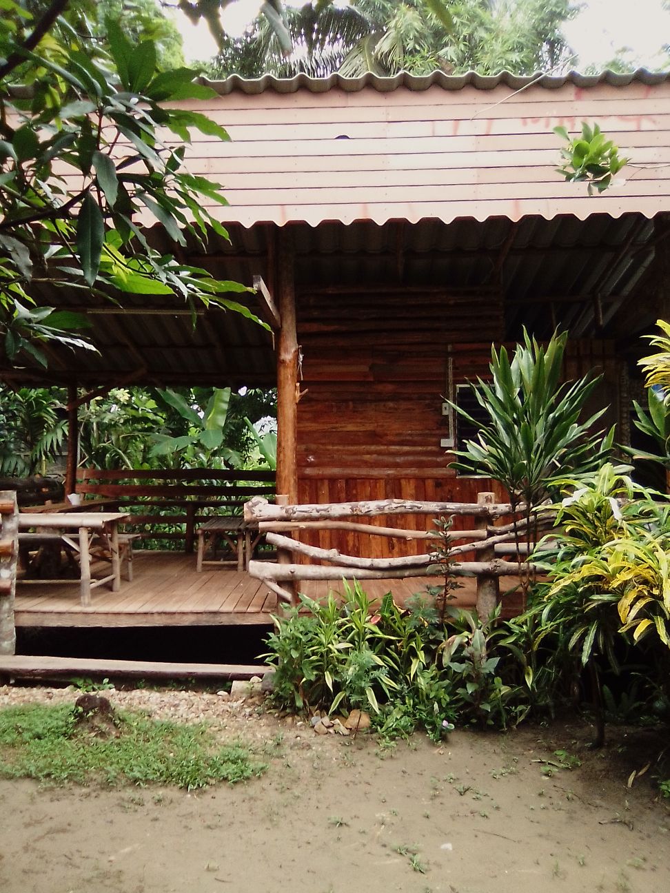 A rustic wooden cabin is surrounded by lush greenery, featuring a spacious outdoor seating area on a raised deck. The structure showcases natural wood finishes, with vibrant plants framing the entrance and providing a serene, harmonious connection to the natural environment.
