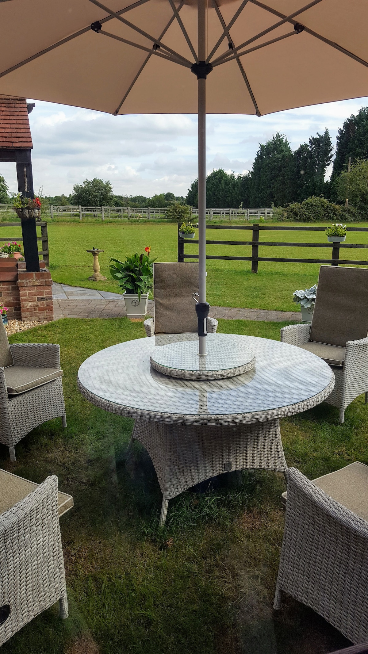 A round outdoor dining table with a central umbrella is surrounded by six wicker chairs on a grassy area. The setting offers views of the surrounding greenery and a wooden fence in the background, contributing to a peaceful outdoor atmosphere.