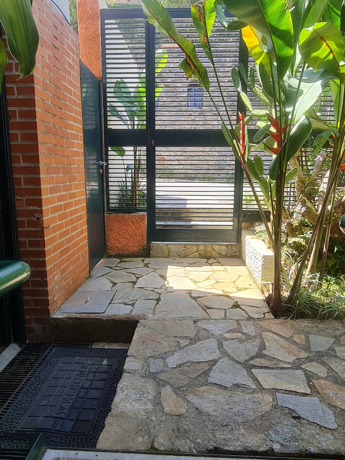 The entryway is framed by lush tropical plants and stone pathways, leading to a gated entrance. Sunlight filters through slatted windows, illuminating the natural stone floor and highlighting the greenery. A textured mat adds an inviting touch to this serene outdoor space.
