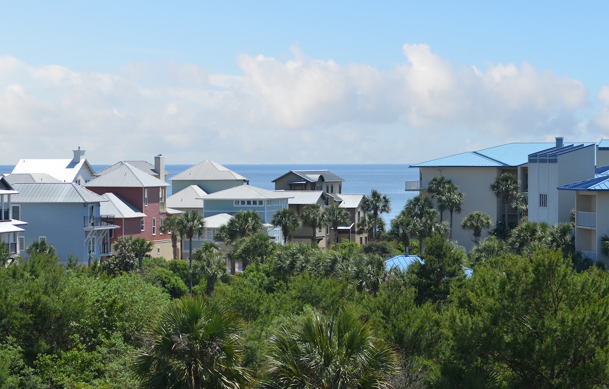 A coastal view showcases nearby buildings with varying architectural styles, framed by lush greenery and palm trees. The horizon reveals the shimmering ocean underneath a bright sky with scattered clouds, creating a serene atmosphere.