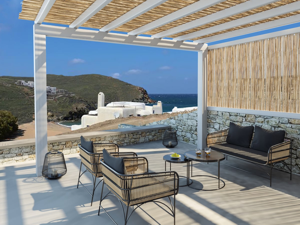 An outdoor seating area features a modern design with two rattan chairs and a sleek sofa, all arranged around a circular table. A view of the coastline and Aegean Sea is visible through a pergola, providing natural light and a refreshing ambiance.