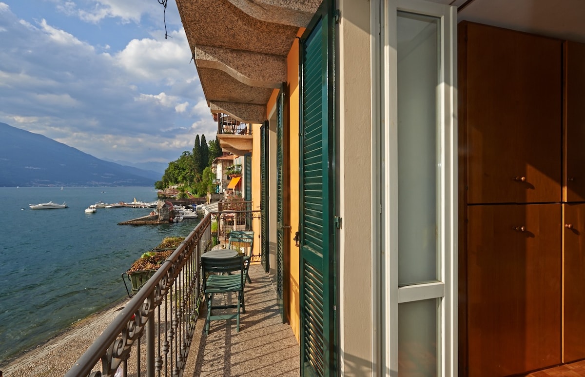 A spacious balcony offers a direct view of Lake Como, featuring outdoor seating and a railing. The shore with pebbles can be seen below, along with boats in the water. Nearby, lush greenery and mountains provide a scenic backdrop.