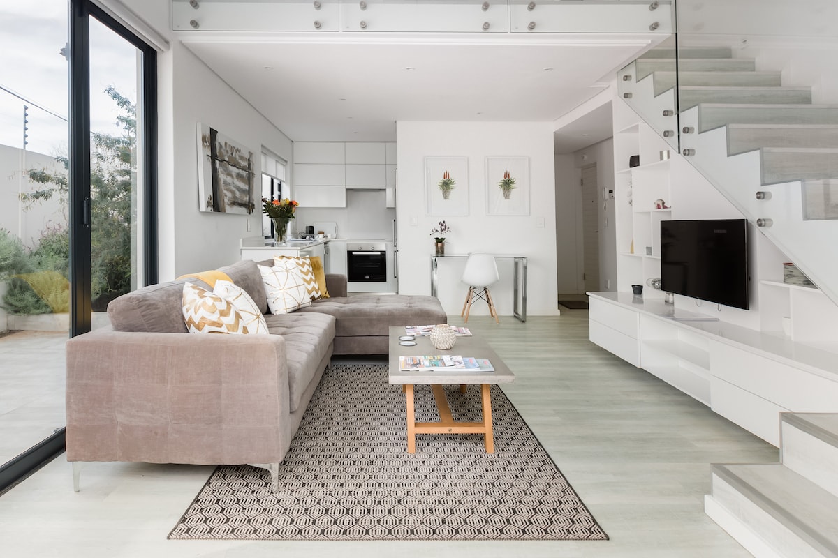A furnished living area features a spacious sectional sofa adorned with decorative pillows. A low coffee table sits on an area rug, complemented by a flat-screen Smart TV mounted on the wall. The open-plan space is bright, with large windows providing views of the garden.