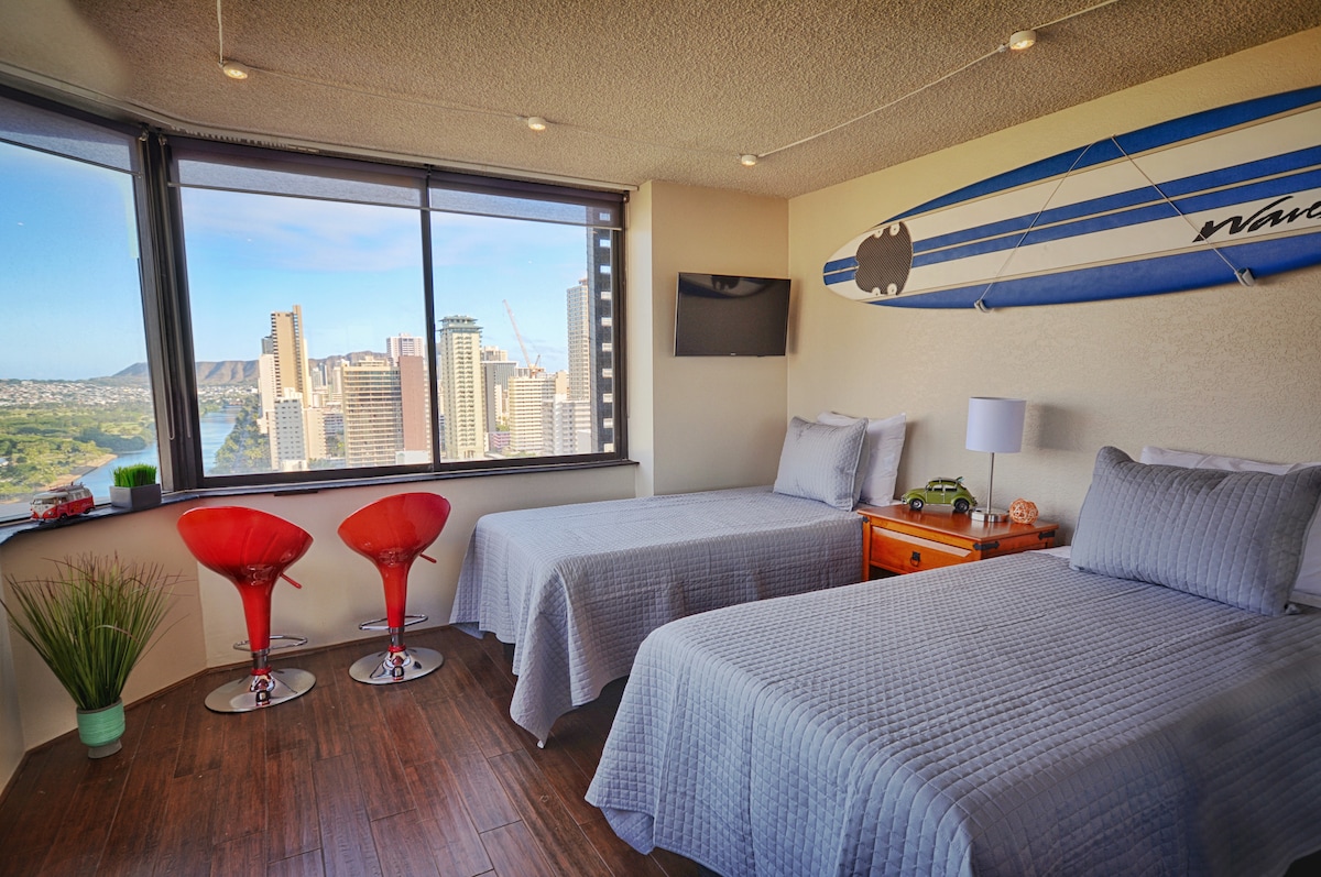 A bright studio space showcases two twin beds with gray bedding, positioned near wrap-around windows. The panoramic city view, including a glimpse of Diamond Head, is framed by the glass. Two contemporary red chairs sit beside a nightstand, adding a modern touch to the room.