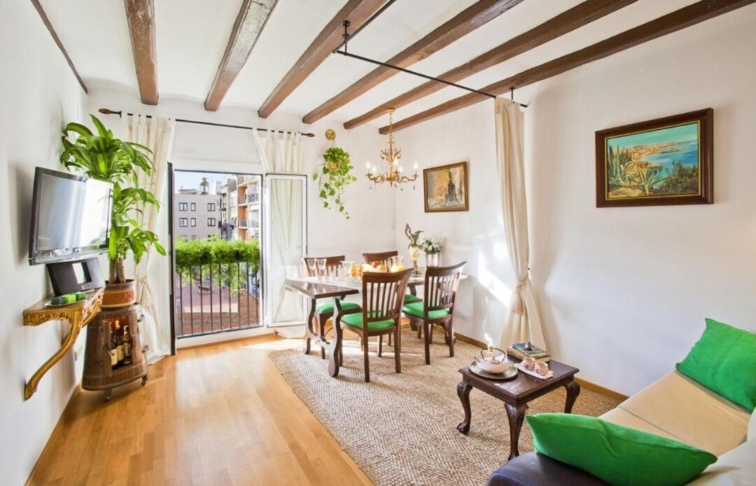 A bright living room showcases wooden beams across the ceiling, complemented by a comfortable sofa and a small coffee table. A dining table with four chairs is visible, along with a TV mounted on the wall. A door leads to a balcony with green views.