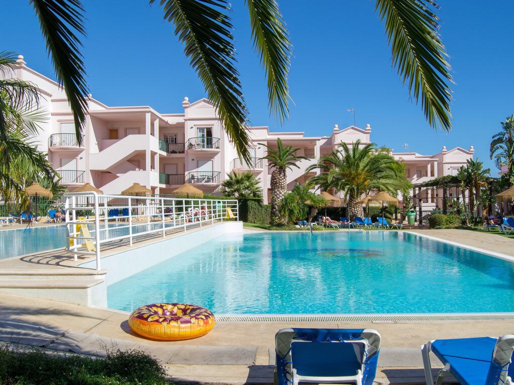 The image showcases a large swimming pool with clear blue water surrounded by a sun terrace. Palm trees and lounge chairs provide shade and relaxation. Brightly colored pool floats are visible, along with a backdrop of a well-maintained apartment complex with balconies and lush landscaping.