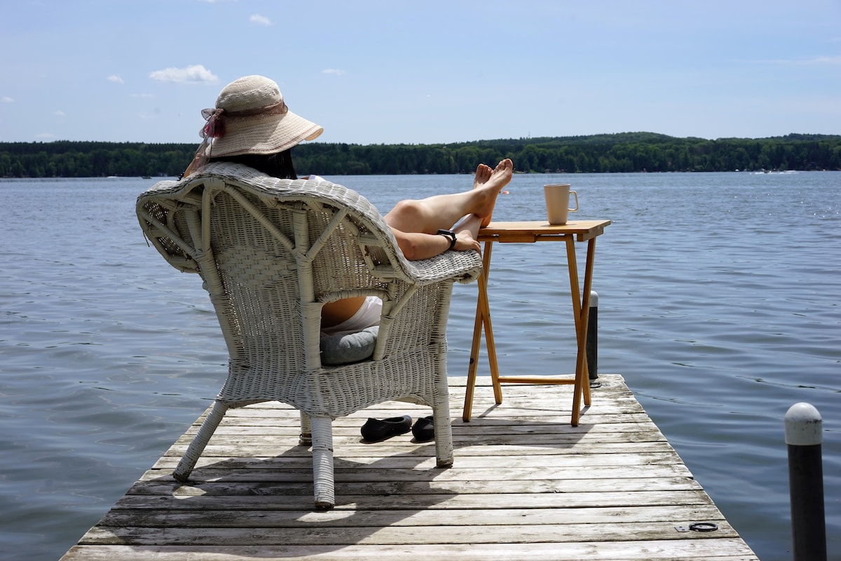 A person is seen relaxing on a wicker chair on a wooden dock extending over a calm lake. A small side table holds a cup, while the serene water reflects the blue sky and greenery in the distance.
