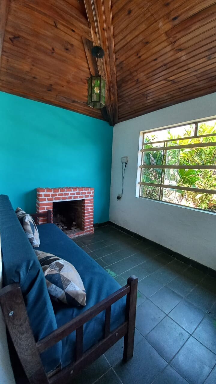 A sitting area features a brick fireplace against a turquoise wall. A wooden bench with decorative pillows is positioned nearby, complemented by a window that offers views of lush greenery outside. The dark tiled floor enhances the space's natural light.