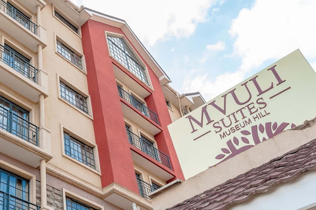 The exterior of the building displays a harmonious blend of cream and red faƧades, complemented by dark-framed windows. A prominent sign reading 'Mvuli Suites Museum Hill' is showcased on the side, indicating the establishment's welcoming identity. The structure features multiple stories with a sloped roof.