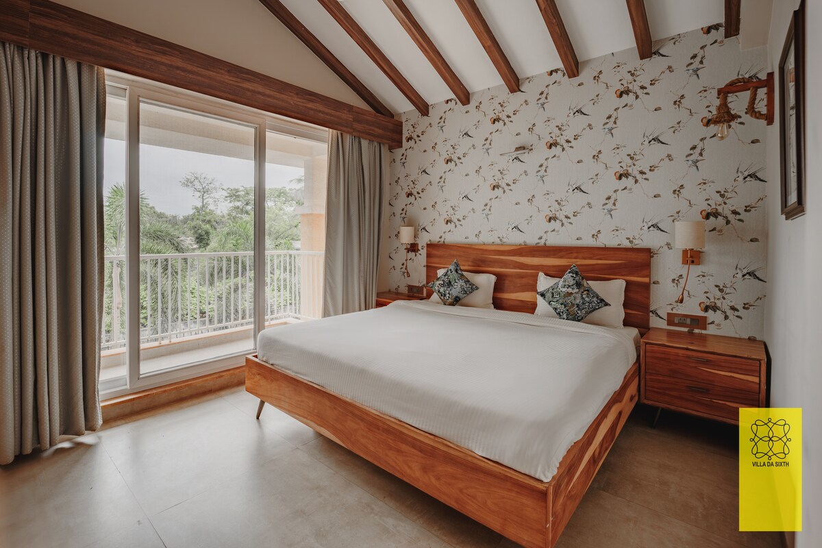 A spacious bedroom is highlighted with wooden accents and a large window offering views of surrounding greenery. The king-size bed is complemented by decorative pillows. Soft curtains frame the window, and bedside tables are positioned on either side of the bed.