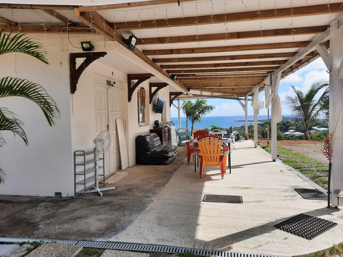 An open terrace area is visible, featuring a seating arrangement with bright, multi-colored chairs. A dark sofa and a ceiling fan are present, with a view of the ocean in the background. The space is shaded by a wooden overhang, creating a relaxed atmosphere.