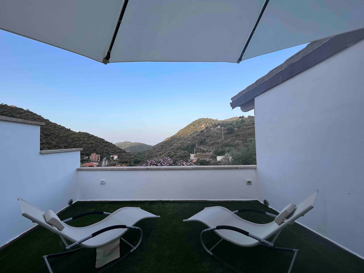 A spacious terrace features two lounge chairs positioned under a large umbrella. The surrounding view encompasses rolling hills and greenery, providing a serene backdrop for relaxation. The terrace is bordered by white walls, enhancing the open-air experience.
