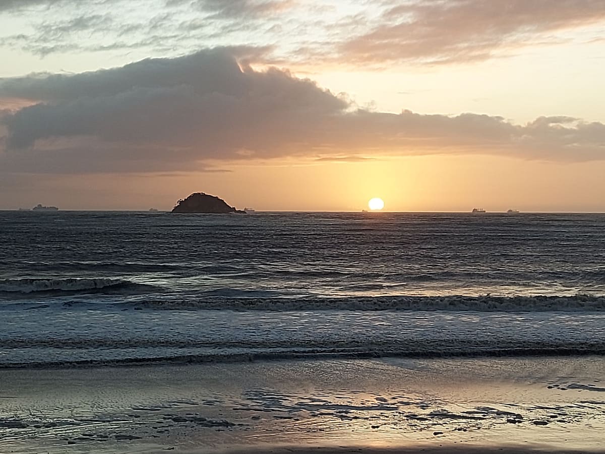A serene view of the ocean at sunset, with the sun descending over the horizon. Gentle waves can be seen lapping against the shore, reflecting soft hues of orange and pink in the sky. A distant island is silhouetted against the fading light.
