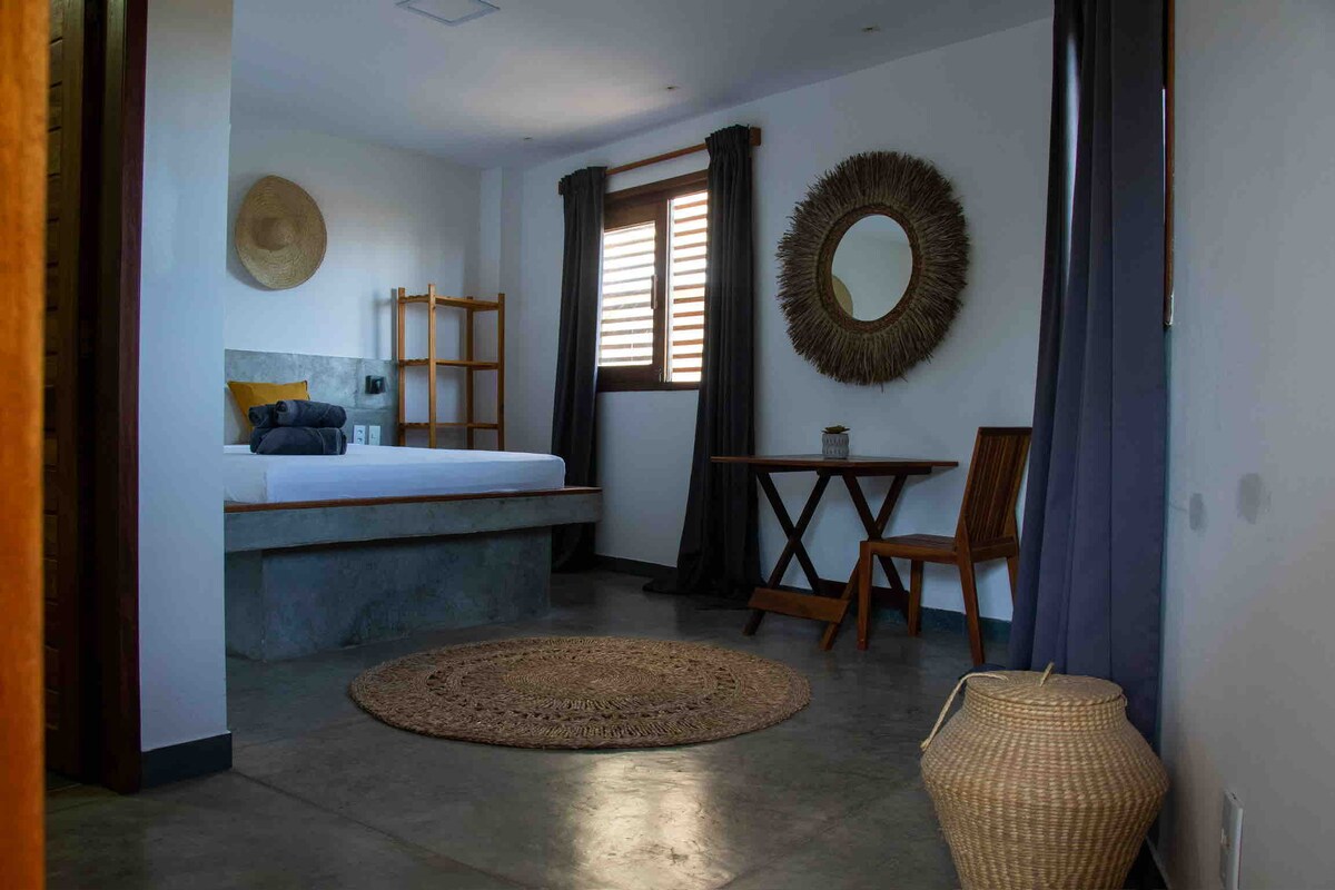 A spacious and modern room features a large bed set against a concrete wall. A round mirror and decorative wall art provide visual interest. A wooden table and chair are positioned near the window, with light filtering through the wooden shutters. A woven rug adds texture to the floor.
