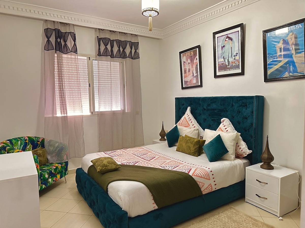 A spacious bedroom is highlighted by a plush bed with decorative pillows and a patterned blanket. The room features large windows covered with sheer curtains, allowing natural light to filter in. Artwork adorns the walls, complementing the room's calming color scheme.