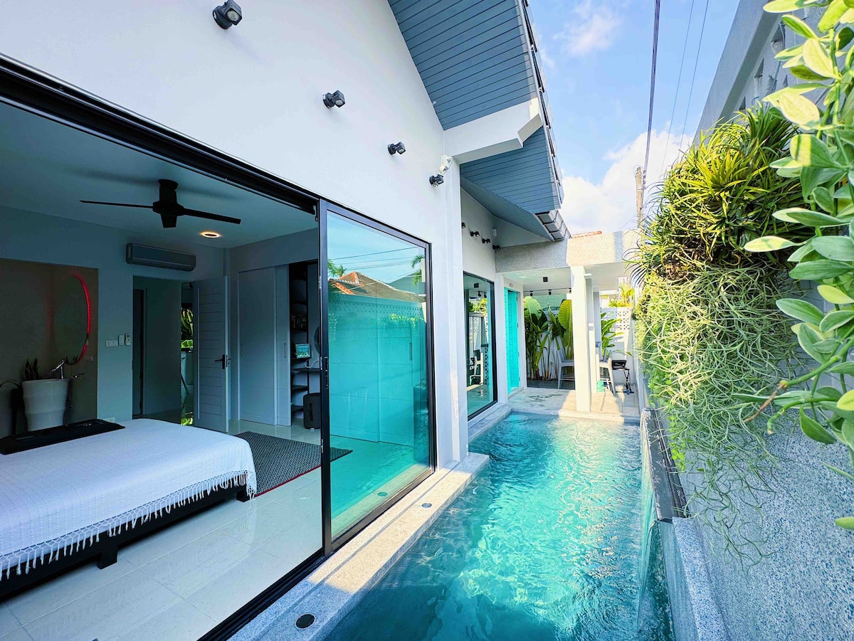 The image captures a modern villa exterior, featuring a private salt-water pool beside a spacious glass-walled living area. Natural light enhances the sleek design, with greenery visibly accentuating the tranquil space. The pool reflects the serene blue sky above, inviting relaxation.