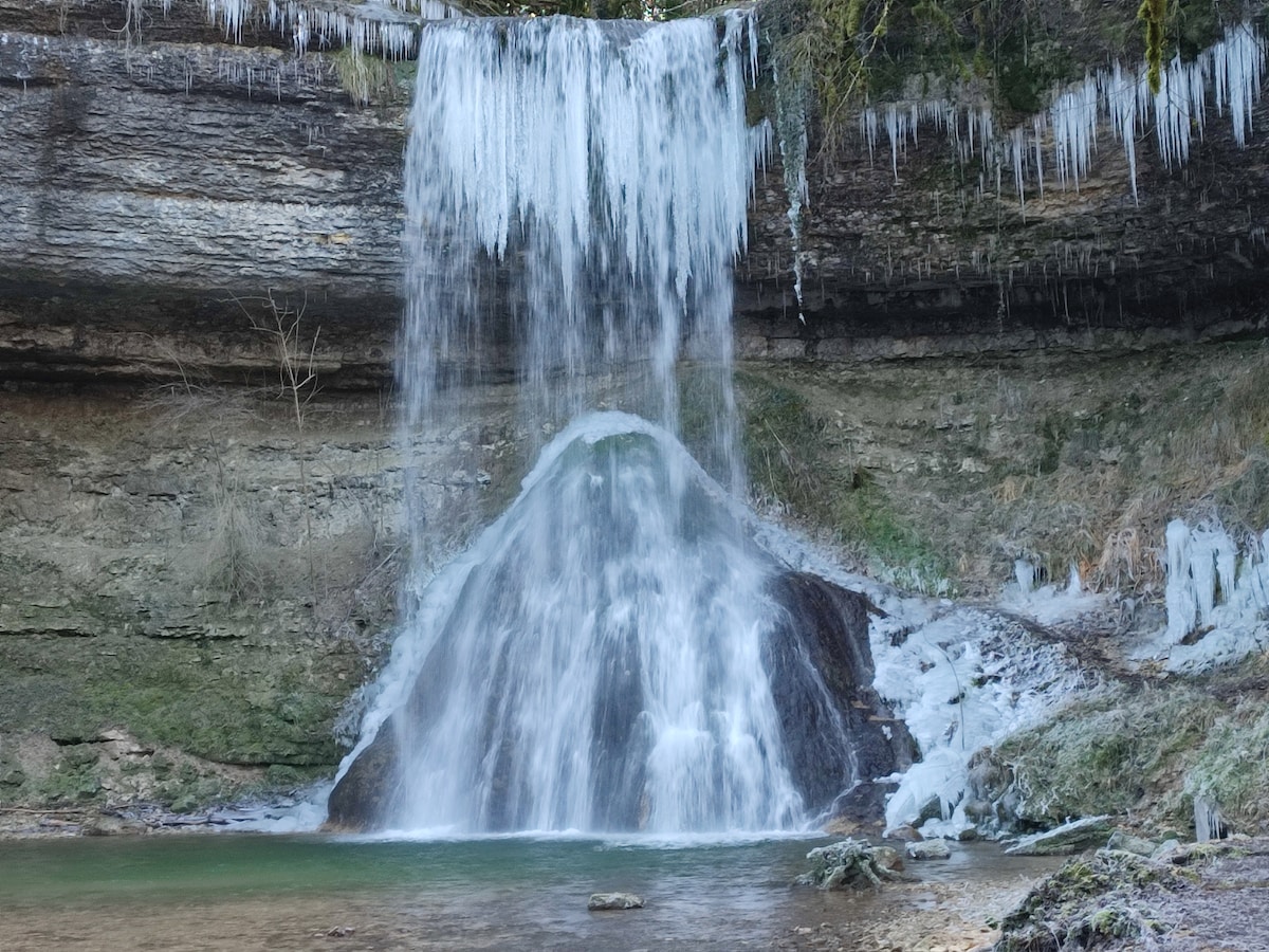 A frozen waterfall cascades down a rocky ledge, surrounded by stone walls. Ice formations hang from the edges, while a small pool collects water at the base. The natural landscape features mossy stones and sparse vegetation, illustrating a serene and tranquil environment.