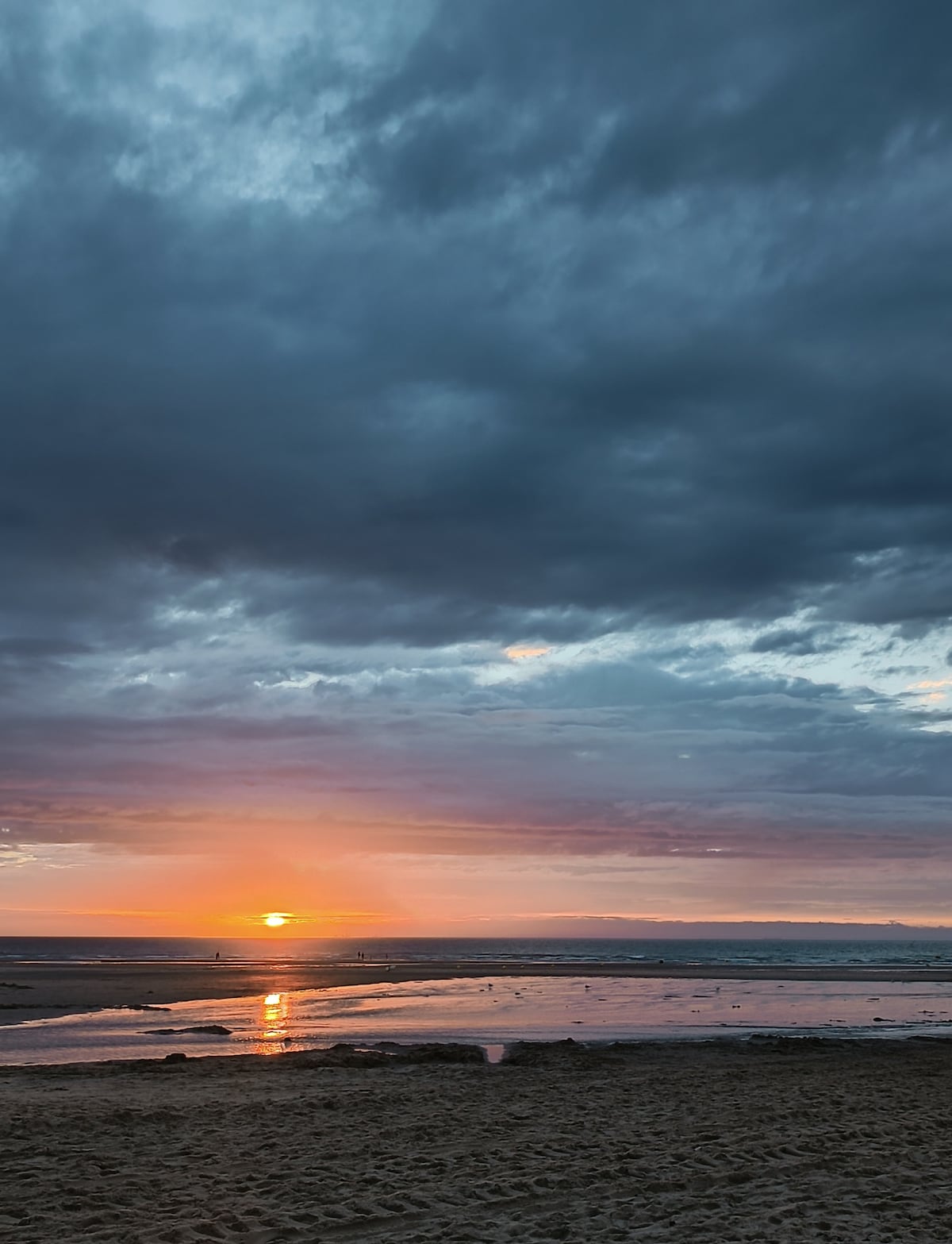 A serene beach scene captures the sunset reflecting over the calm waters. The sky features a blend of dark clouds and warm hues, while the wet sand creates a glistening effect. The horizon showcases the sun dipping below, contributing to a tranquil atmosphere.
