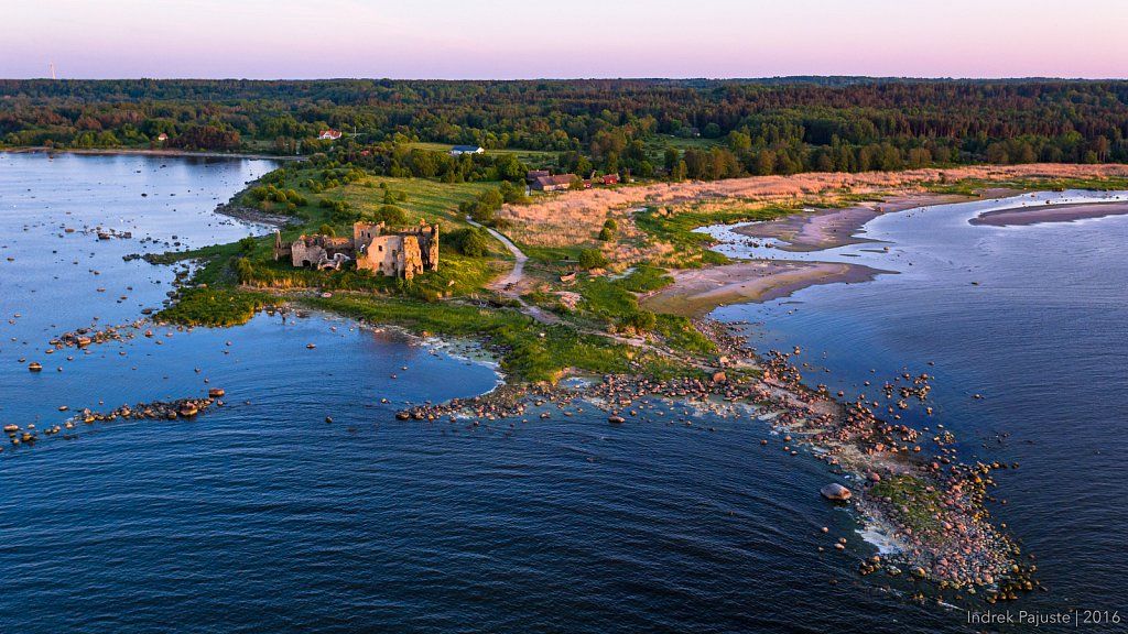 Aerial view of a coastal landscape featuring an old, partially ruined structure near the water's edge. The shoreline is bordered by rocks and sand, leading to a green area with trees in the background. A serene body of water reflects the pale hues of sunset.