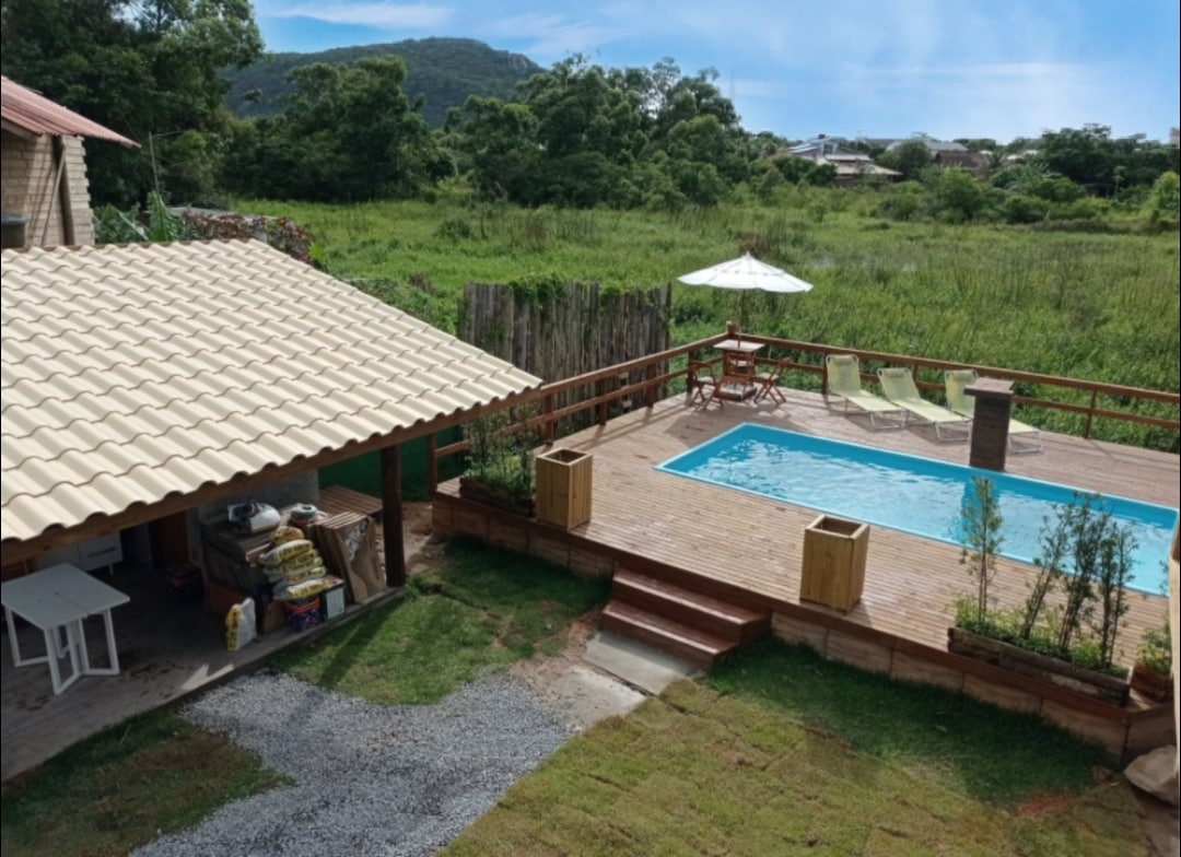 A wooden deck surrounds a rectangular pool, with sun loungers positioned nearby. A shaded seating area is offered under a tiled roof, with a view of the lush greenery and hills in the distance. The space reflects a serene, nature-filled environment.