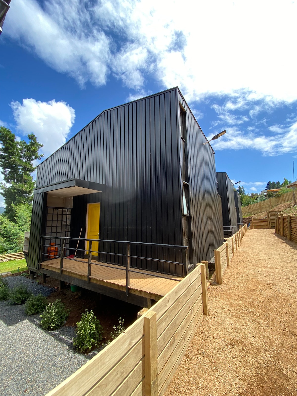 A modern two-story cabin is depicted, characterized by its black facade and geometric design. A wooden walkway leads to the entrance, surrounded by gravel pathways and greenery. The background features a clear blue sky dotted with fluffy white clouds.