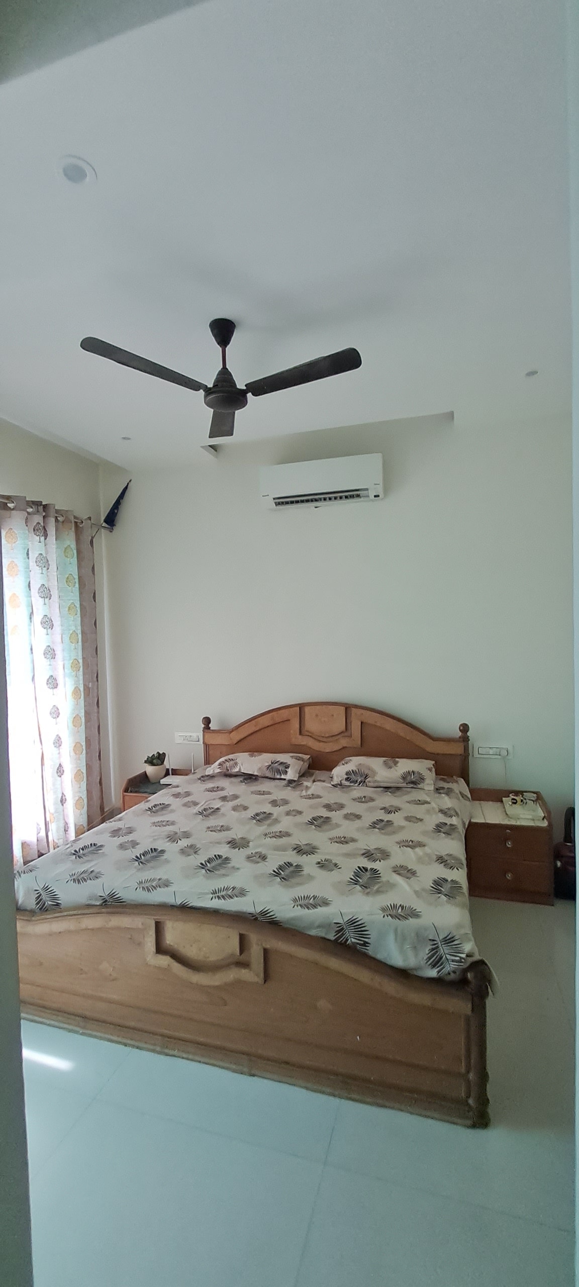A well-lit bedroom features a spacious bed with a patterned blanket. A ceiling fan is positioned above, and an air conditioning unit is mounted on the wall. Natural light enters through a window adorned with patterned curtains, complementing light-colored walls and flooring.