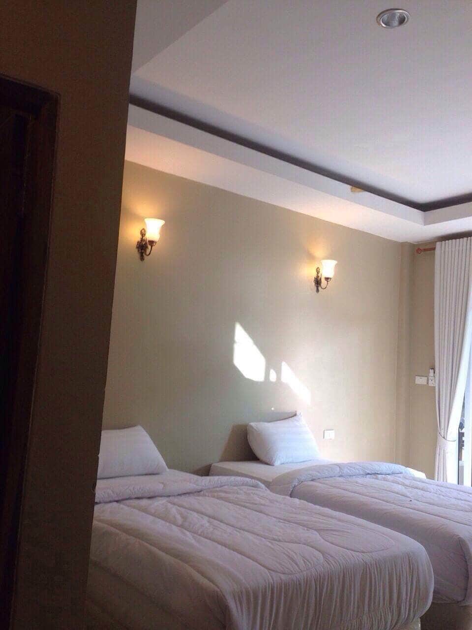 A well-lit room features two spacious beds with white linens, positioned against a soft green wall. Wall sconces provide gentle lighting, and natural light enters through sheer curtains on a nearby window, creating a calm and restful environment.
