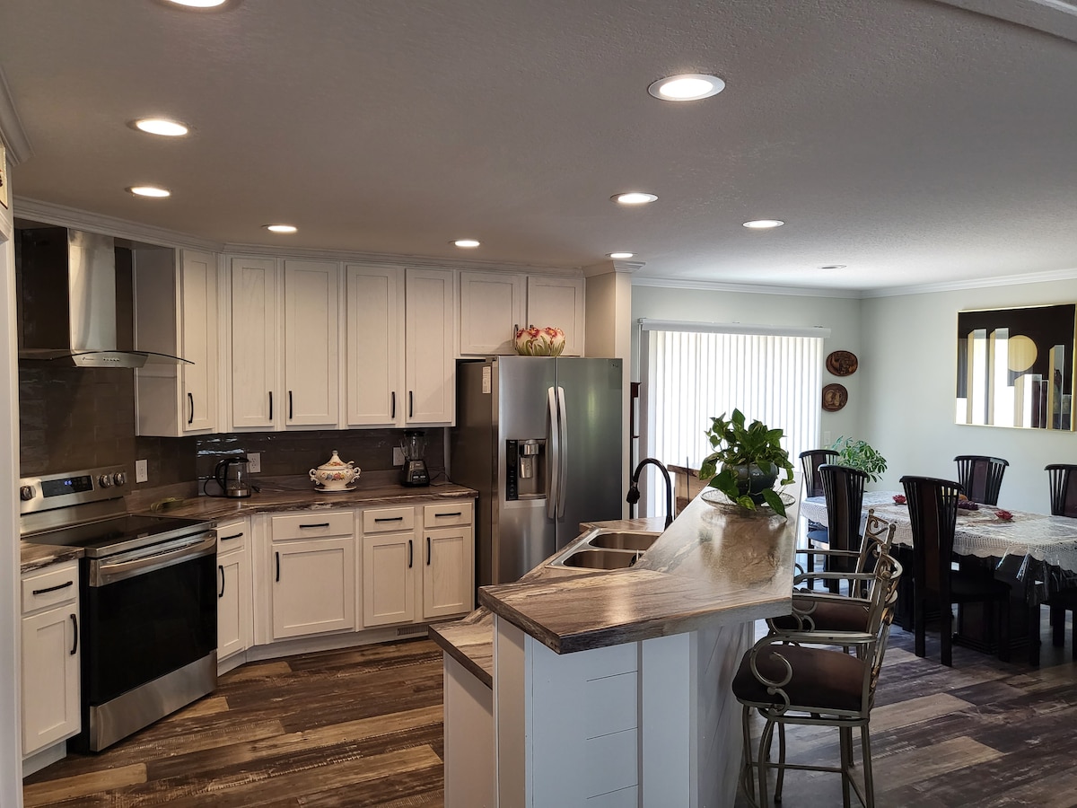 A spacious kitchen features modern stainless-steel appliances, white cabinetry, and a light-colored counter. Natural light floods the room from a large window, illuminating a dining area with a table surrounded by dark chairs. Decorative items and greenery add a touch of warmth.