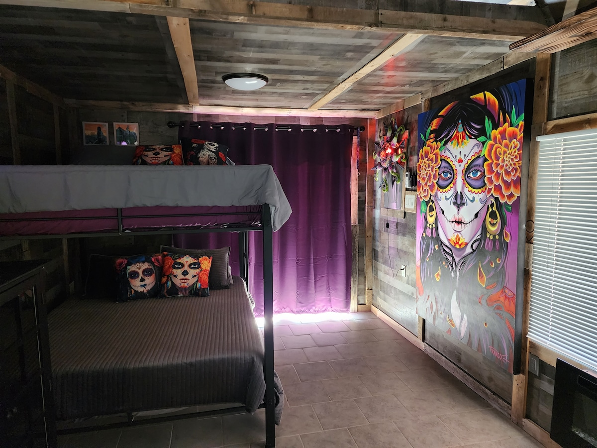 A cozy room features a bunk bed with a bottom double-sized bed and a top single bed. Brightly colored wall art decorates one wall, while purple curtains frame a window, allowing natural light to fill the space. Neutral-toned bedding complements the rustic wood walls.