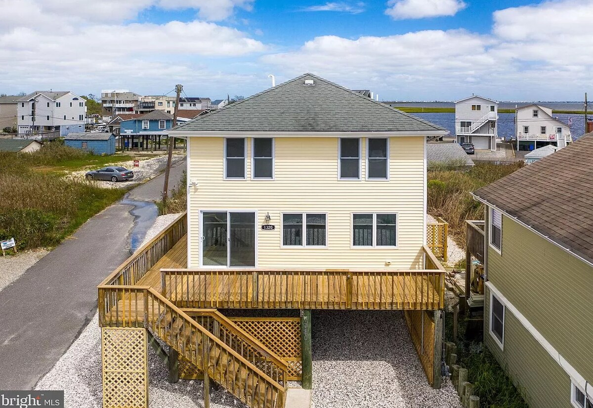The exterior of a two-story yellow house is showcased against a partly cloudy sky. A wooden staircase leads to a wraparound deck, surrounded by gravel landscaping. Nearby homes and the water can be seen in the background, highlighting the coastal setting.