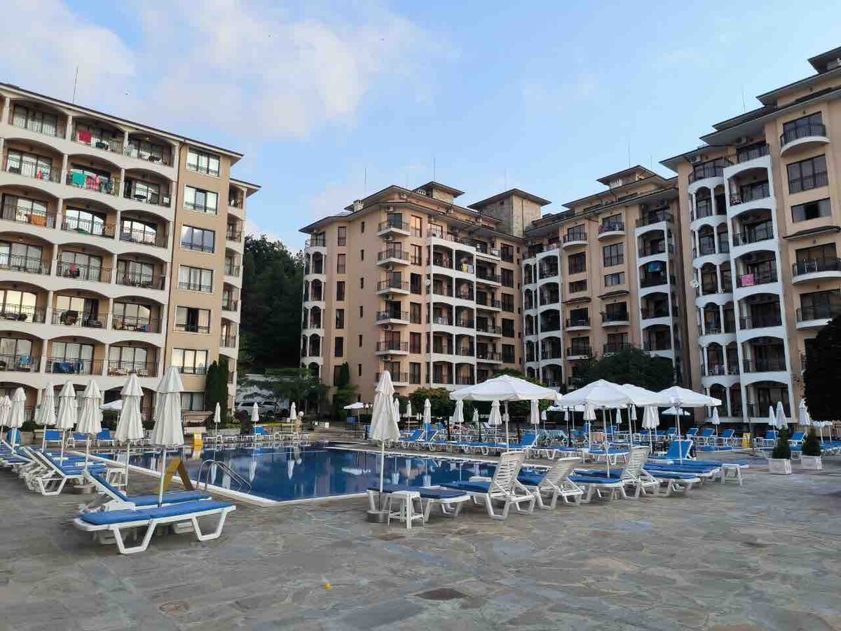 The image captures a spacious outdoor swimming pool surrounded by sun loungers and umbrellas. The buildings, featuring multiple balconies, provide a backdrop to the pool area. Clear skies enhance the inviting atmosphere, suitable for relaxation and leisure.