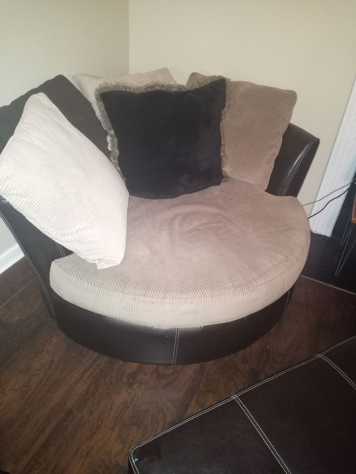 A round, plush chair is positioned in the corner, featuring soft beige upholstery and two large pillows: one in a light texture and the other in a solid black. The chair's base is covered in dark material, complementing the warm wooden floor.
