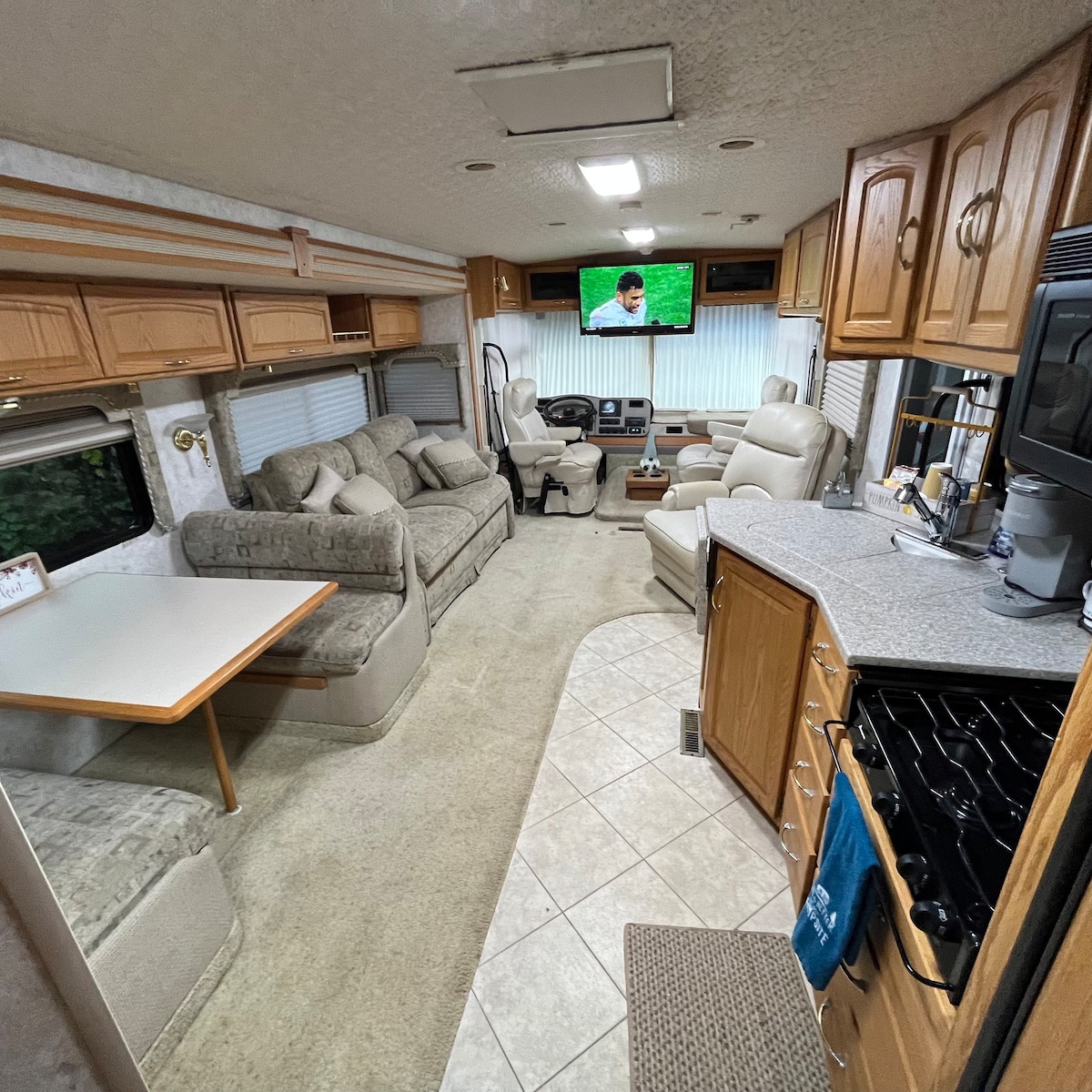 The living space within the motorhome features soft leather seating, a reclining couch, and a table with four chairs. A 40-inch TV is mounted on the wall, while the kitchen area showcases ample counter space and a propane stove. Natural light is provided by nearby windows.