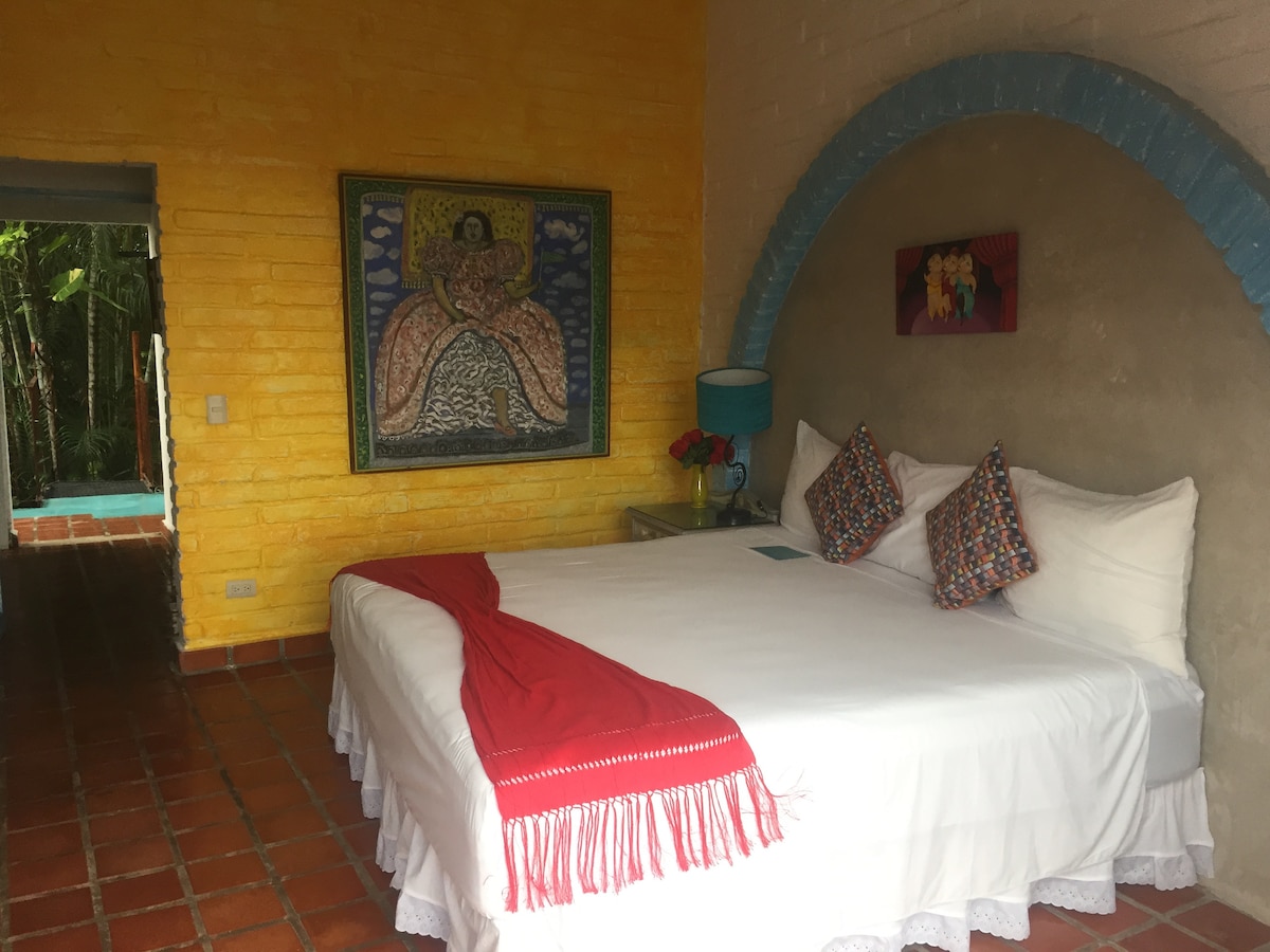 A spacious bed is presented with white linens and decorative pillows, set against bright yellow walls. Unique artwork adorns the wall behind the bed, while a blue arch adds architectural interest. Natural light fills the room through an adjacent door leading to an outdoor area.