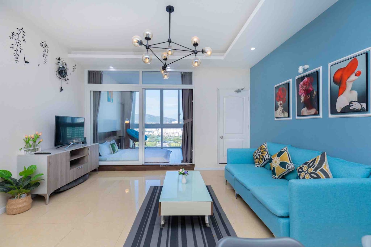 A bright living area features a turquoise sofa adorned with patterned cushions. A modern coffee table sits atop a striped rug, with a flat-screen TV mounted nearby. Natural light filters through large glass doors that open to a view, and art pieces are displayed on the walls.