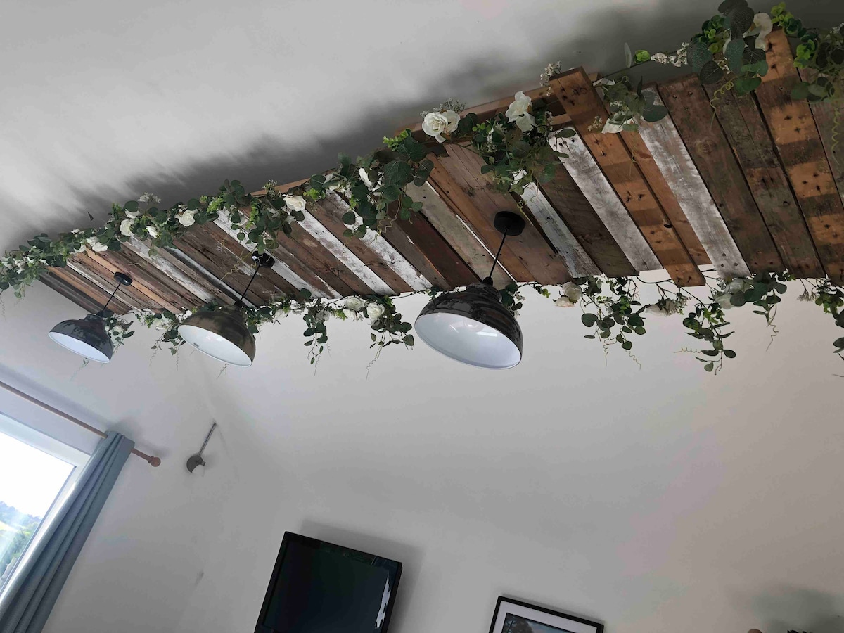 A unique ceiling design features wooden planks and greenery, creating a natural ambiance. Three industrial-style lights hang from the ceiling, offering illumination. A television and framed artwork are visible on the wall below, contributing to the overall aesthetic of the space.