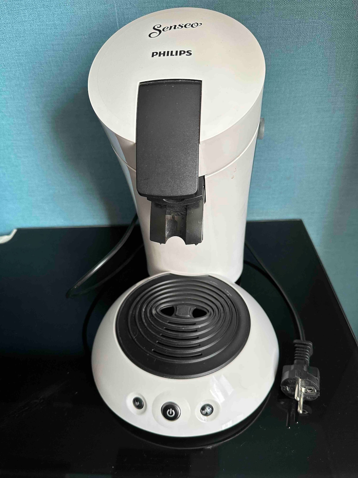 A coffee machine is placed on a dark countertop. The unit is white, featuring a prominent brewing spout and a circular base with control buttons. A power cord is seen plugged in beside it, with a solid blue wall in the background.