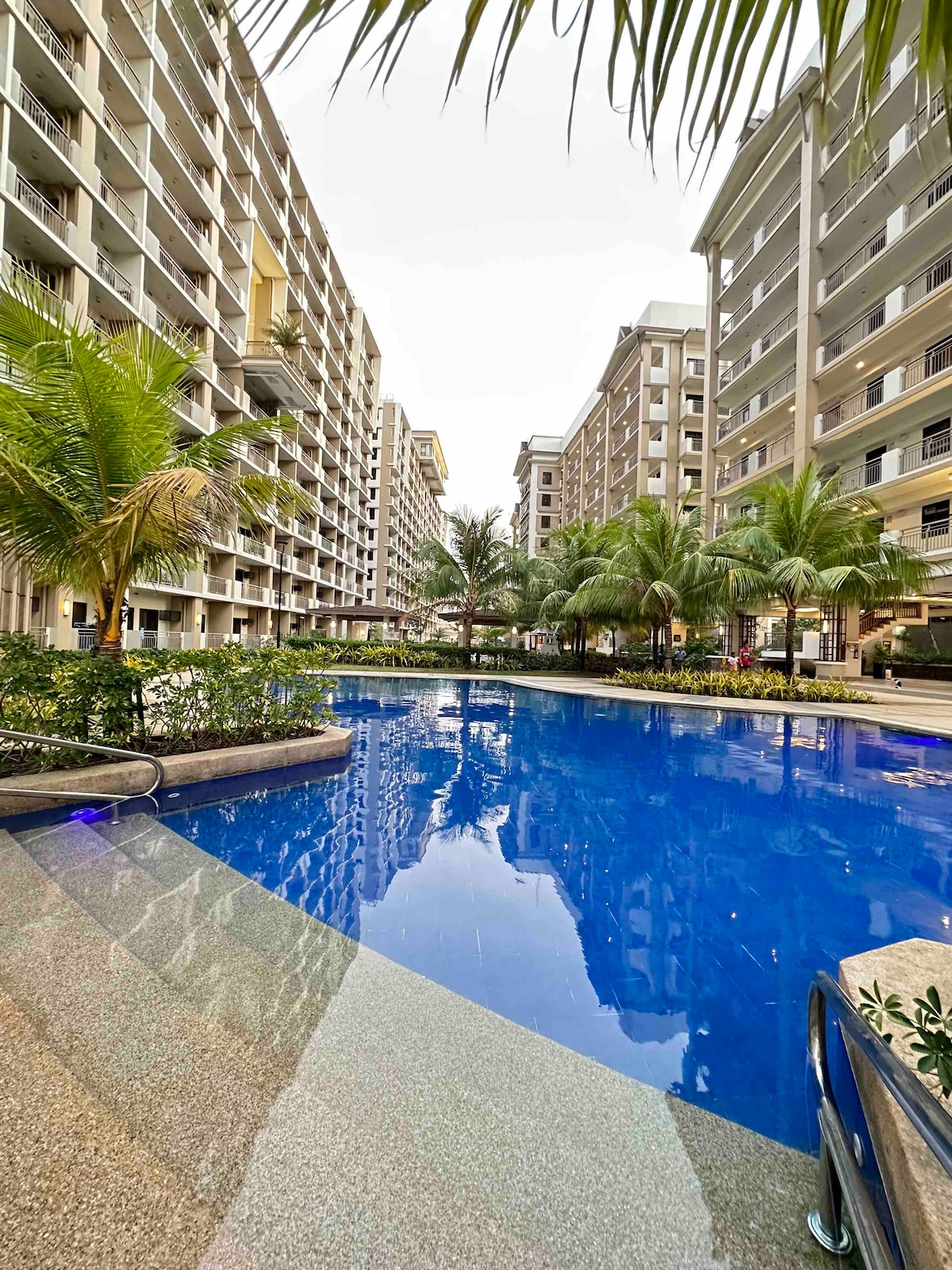 A clear view of the outdoor swimming pool is presented, surrounded by palm trees and landscaped gardens. The pool reflects the surrounding buildings and sky, offering a serene ambiance within the resort-inspired community.