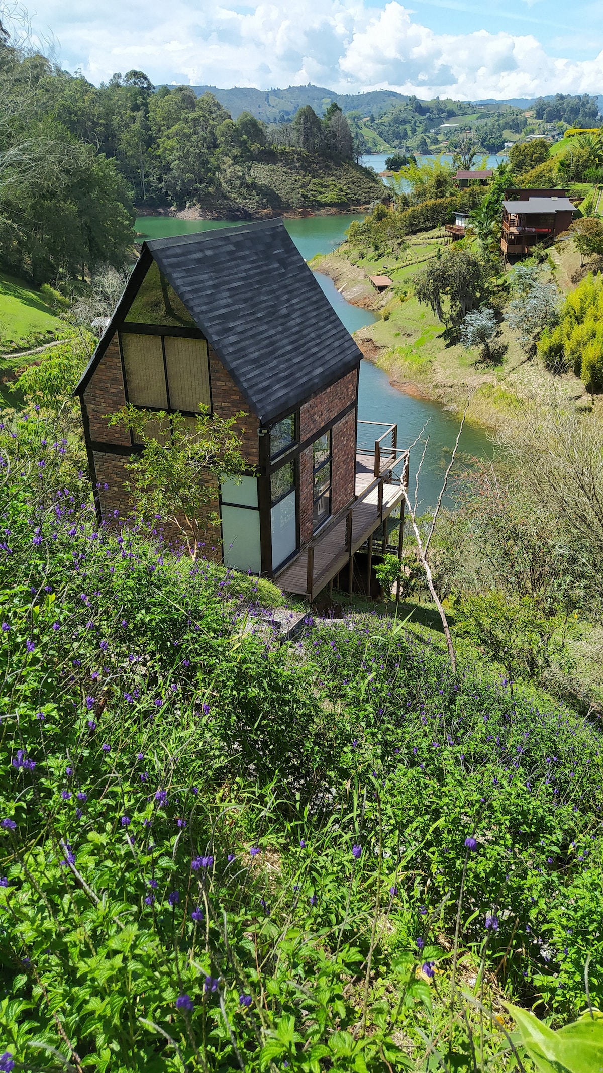 The chalet is positioned on a hillside with a view of the serene waters of the El Peñol-Guatapé reservoir. Lush greenery surrounds the cabin, while nearby trails and vibrant flowers complement the natural landscape.