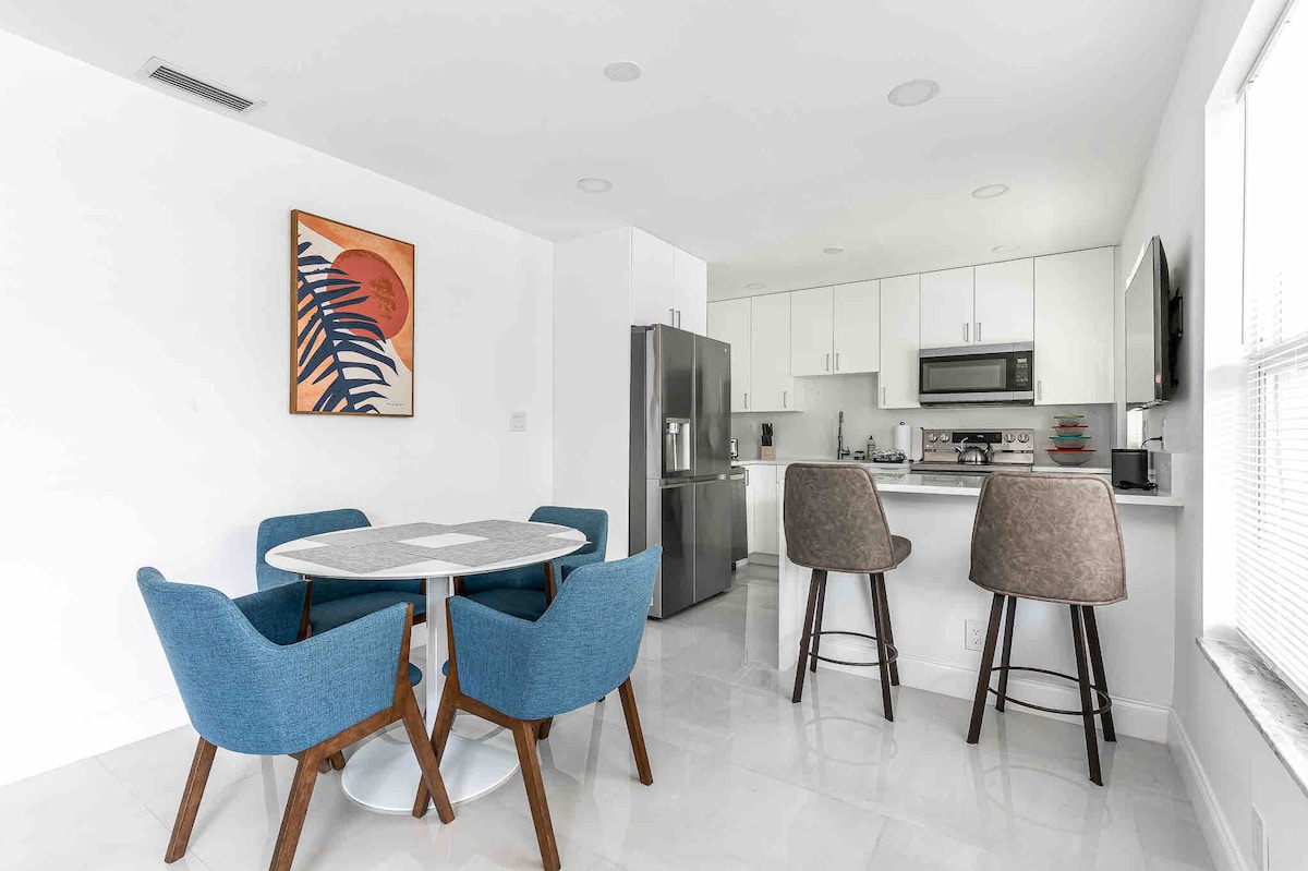 An open-plan kitchen and dining area is depicted, featuring a round dining table surrounded by four blue chairs. A modern kitchen setup is visible, equipped with stainless steel appliances and sleek cabinetry. Natural light enters through a window, enhancing the clean, neutral color scheme.
