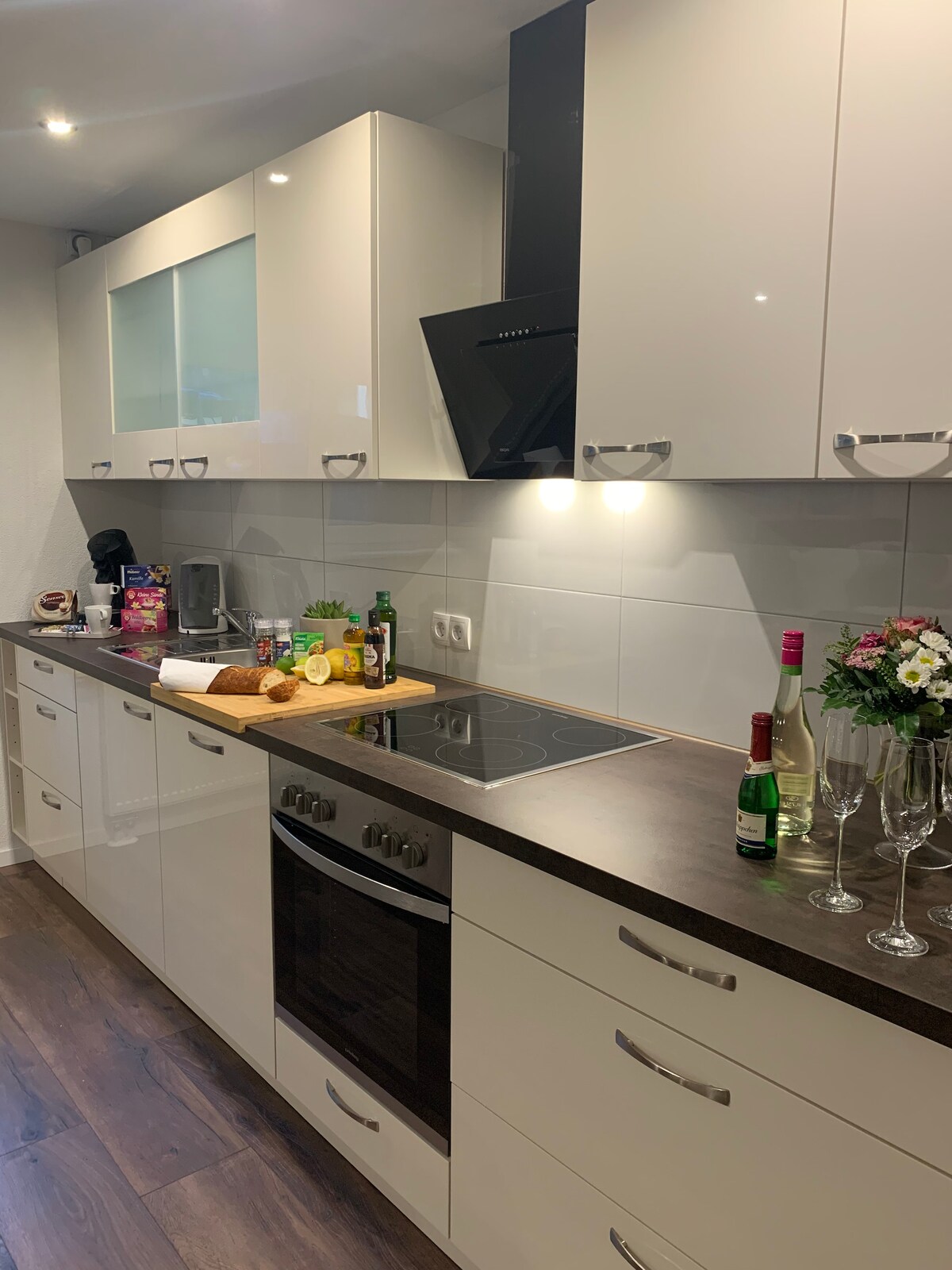A modern kitchen is displayed with sleek white cabinetry and a dark countertop. A built-in oven and stovetop are visible, and a small selection of drinks and snacks are arranged on the countertop. Light illuminates the work area, enhancing the clean, functional design.