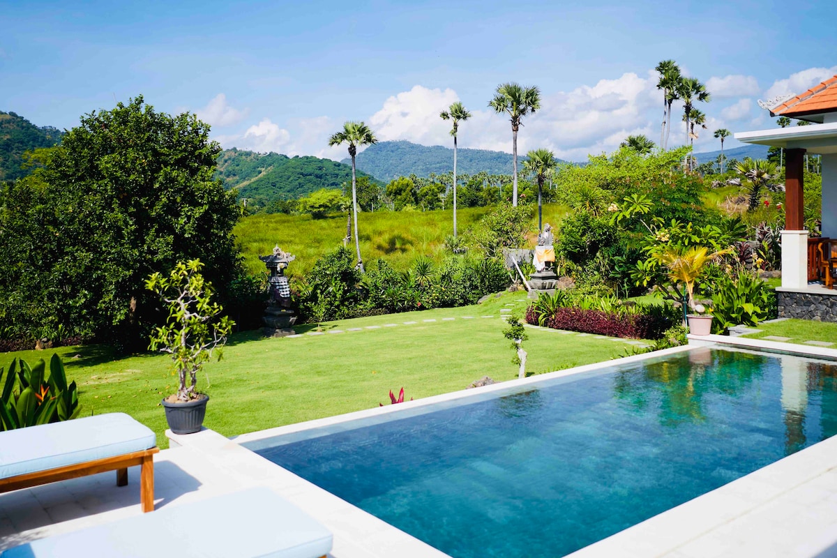 A tranquil private pool reflects the clear blue sky, surrounded by a manicured lawn and lush greenery. Palm trees and ornamental plants frame the space, while distant hills provide a scenic backdrop. The area features lounge chairs for relaxation under the sun.