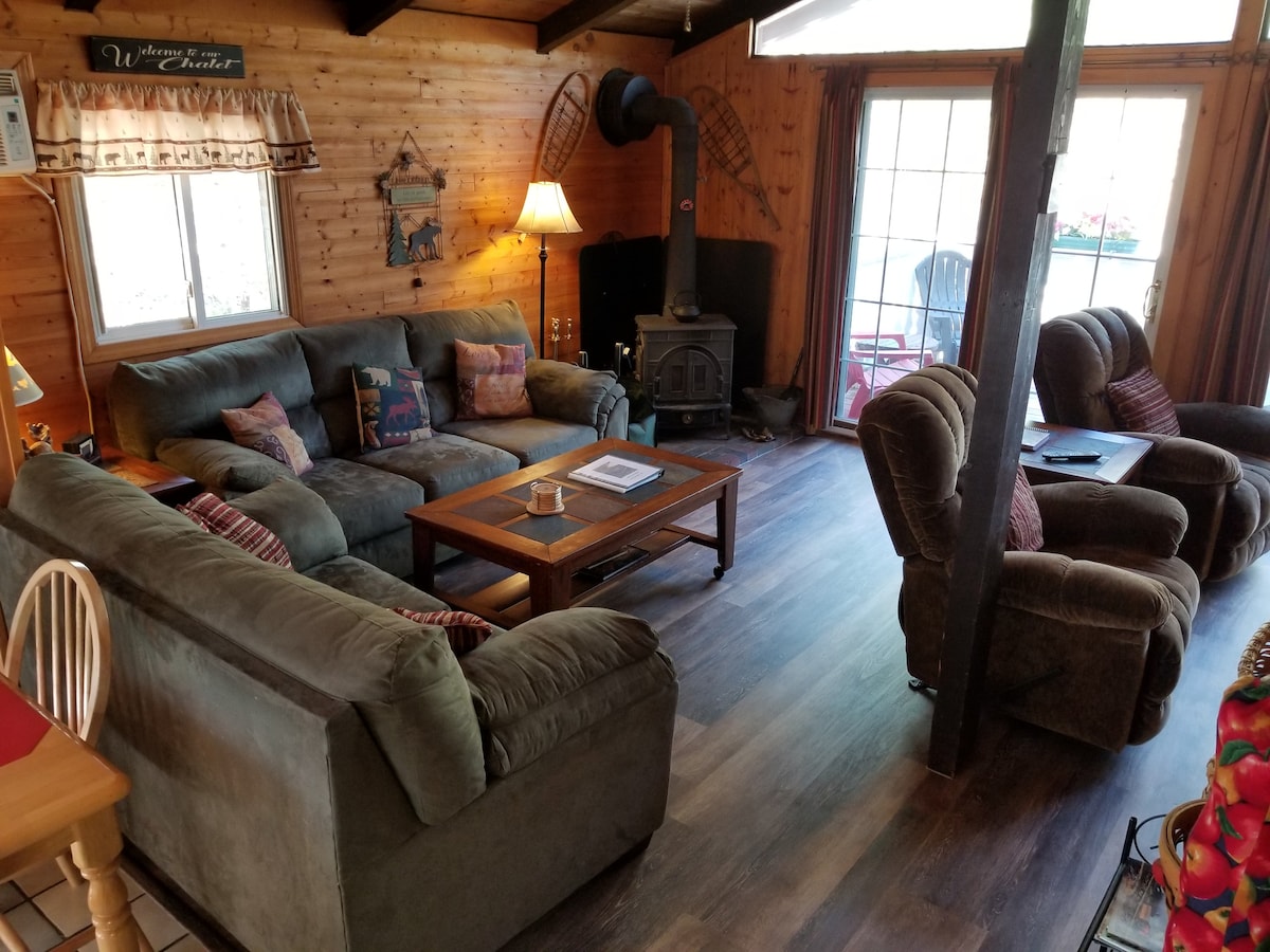An open living area features a large, comfortable sectional sofa and two armchairs, inviting relaxation. A wooden coffee table is centered in the space, with natural light streaming in through the windows. A wood-burning stove adds warmth, while wooden paneling enhances the cozy setting.