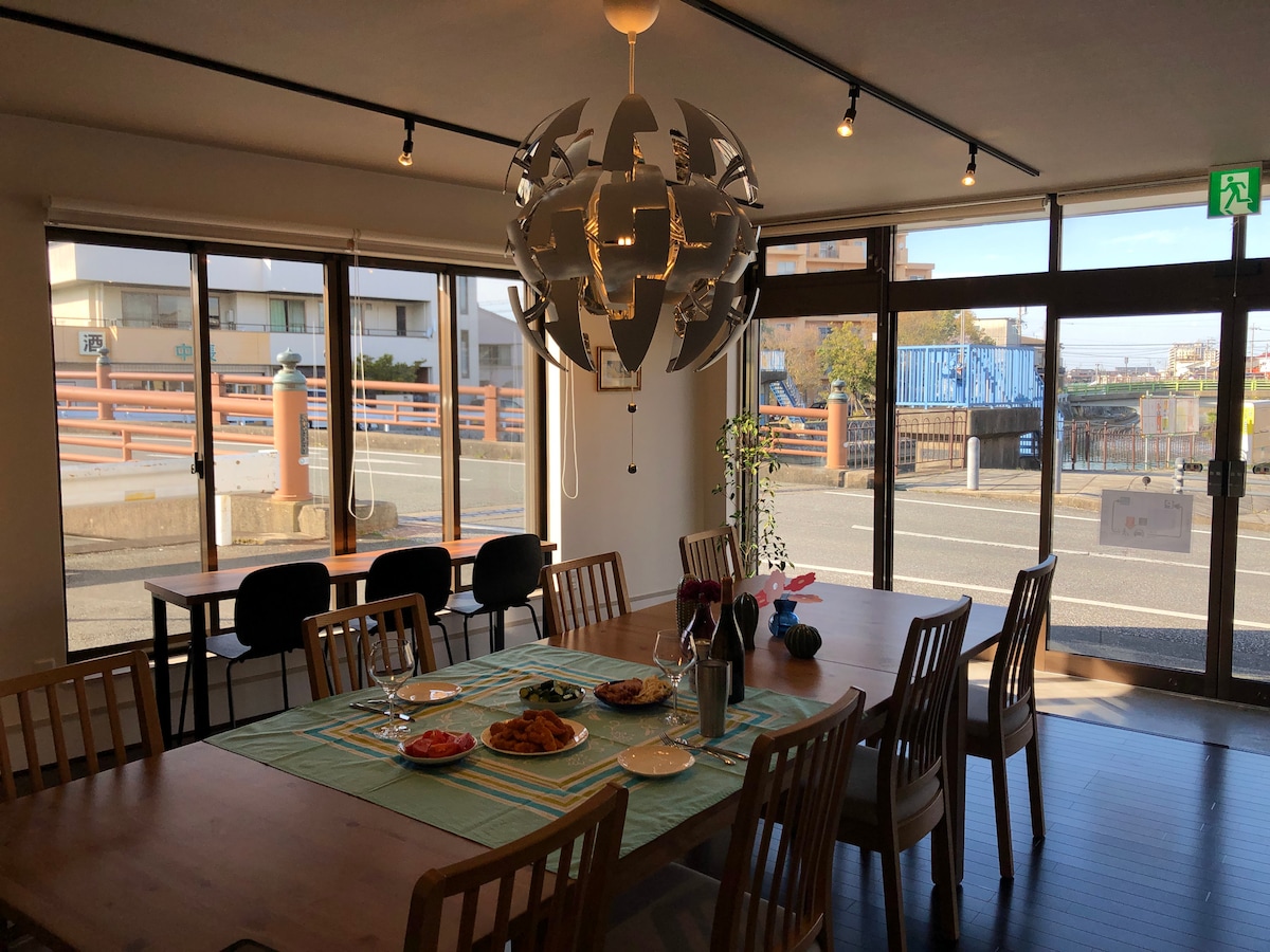 A communal dining area features wooden tables and chairs arranged around a large table set with plates and dishes. A modern light fixture hangs above, and large windows offer abundant natural light and views of the street outside.