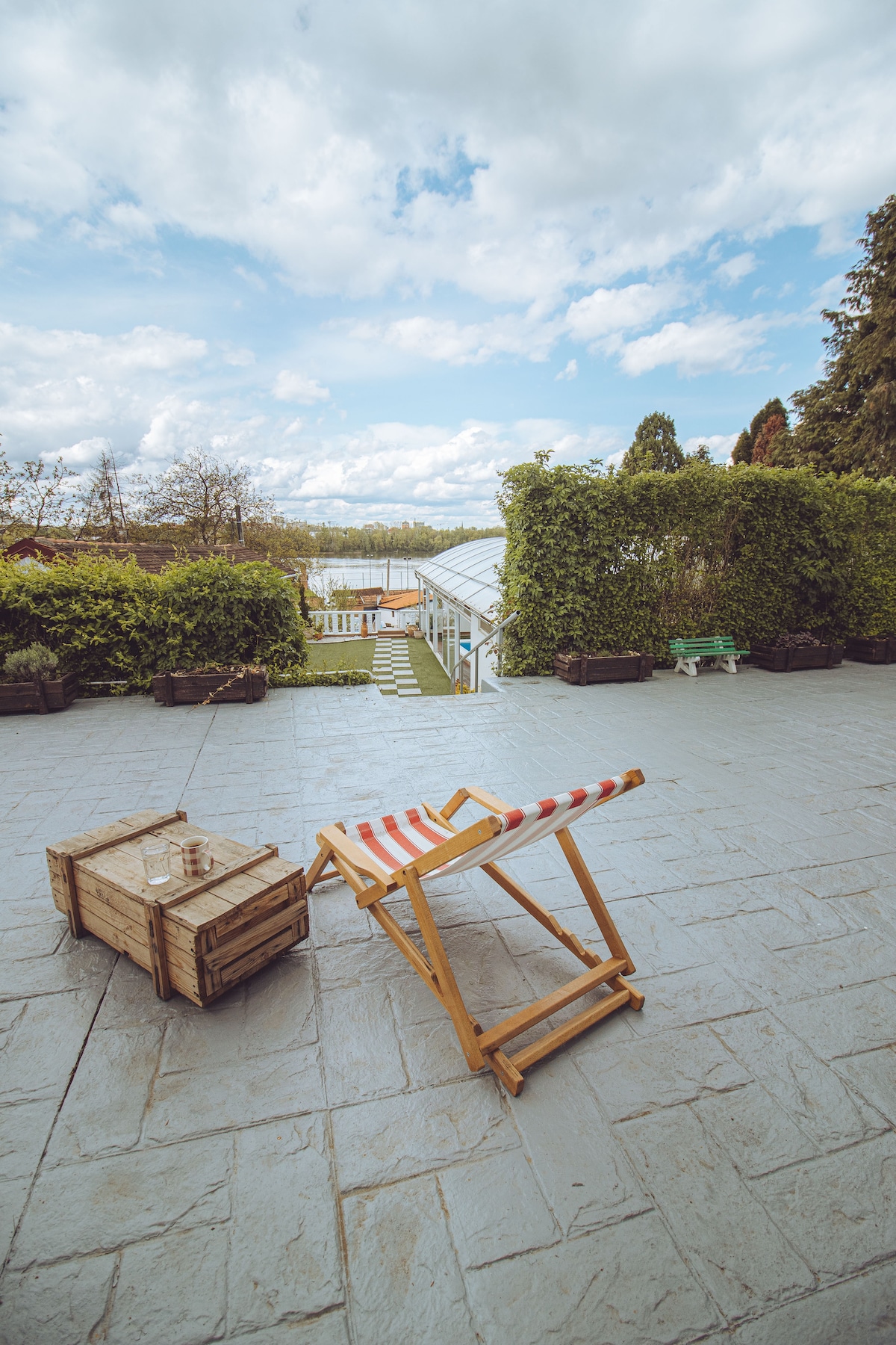 A spacious outdoor area is illustrated with a stone patio, featuring a wooden deck chair with red and white stripes. In the background, lush green hedges provide privacy, while steps lead towards a garden space. A wooden crate holds a glass, adding a touch of charm.