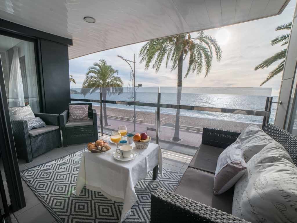 A balcony offers coastal views, with palm trees swaying gently in the breeze. Comfortable seating surrounds a small table set with beverages and fresh fruits. A patterned rug adds texture to the space, creating a relaxing spot to enjoy the surrounding scenery.